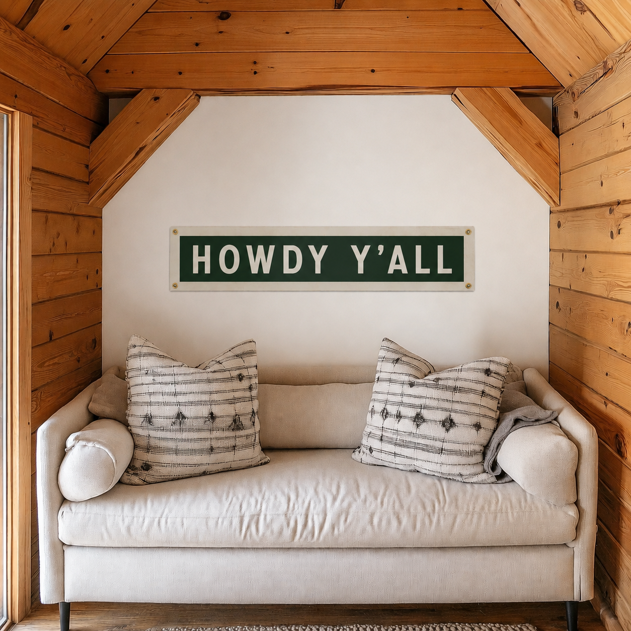 A rustic green and white metal sign reading 'HOWDY Y'ALL' is mounted on a white wall above a cream-colored sofa in a cozy, wood-paneled room.