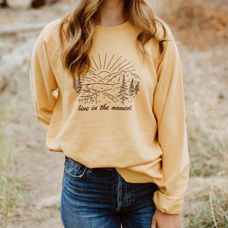 Yellow pullover. Mountain landscape in black linework, with text "live in the moment" under graphic.