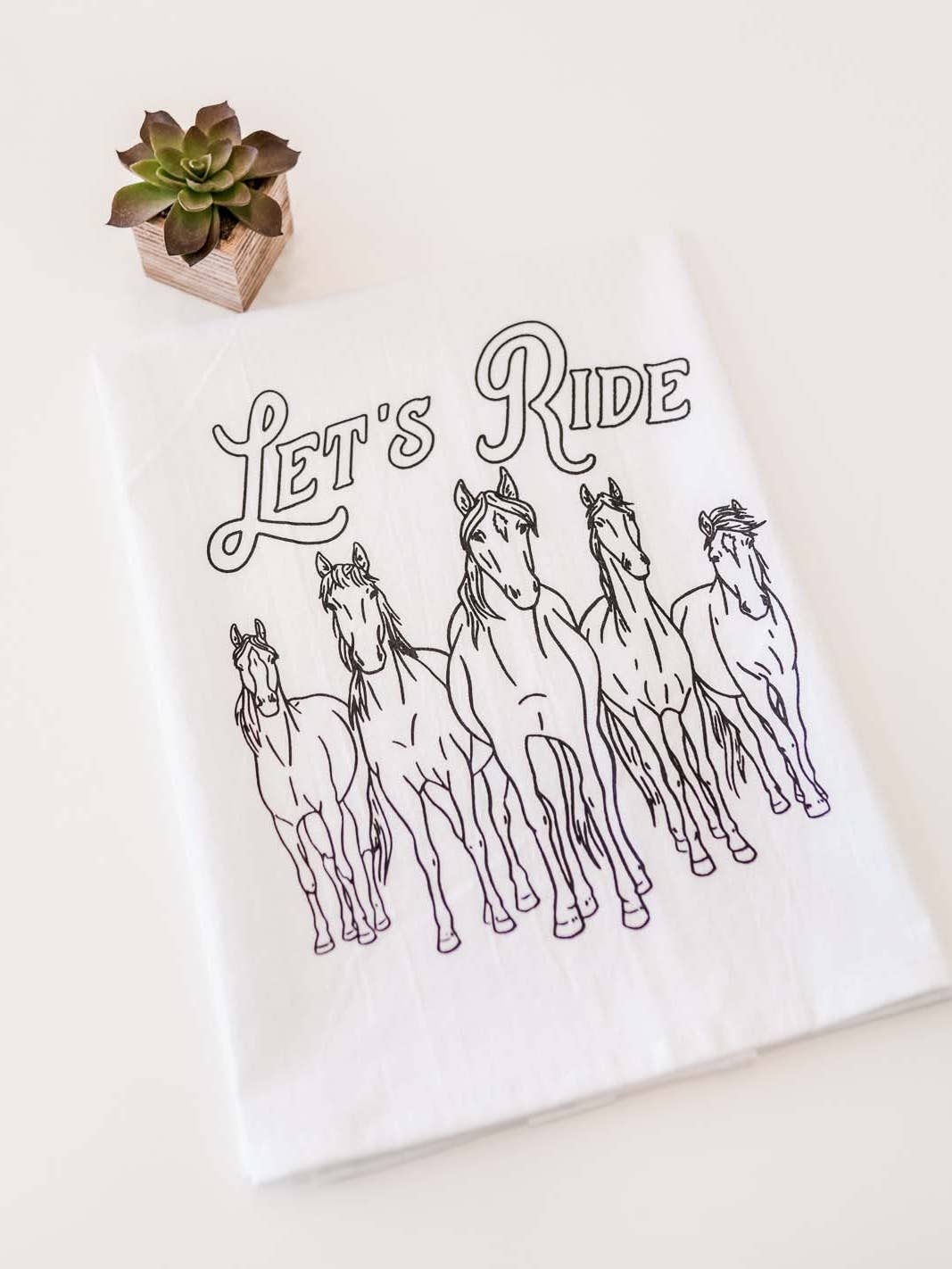 White tea towel with "let's ride" in black lettering. 5 wild horses in black underneath